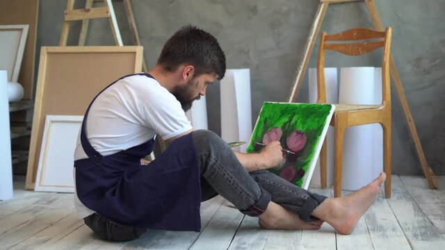 a bearded charismatic Caucasian man sitting barefoot on a wooden floor in an art workshop dressed in a blue apron holding a palette with paints in his hands draws a picture with peonies flowers on a