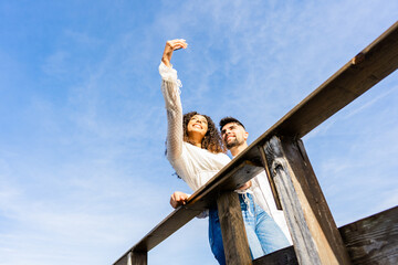 Unusual low angle view shot of multiracial couple on a wooden beach bridge taking a selfie in vacation at sea ocean resort. Large sky copy space ideal for banner or ads. Hispanic girl using smartphone