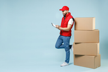Full size body length delivery guy employee man in red cap white T-shirt vest uniform work as dealer courier hold blank cardboard box isolated on pastel blue color background studio. Service concept.