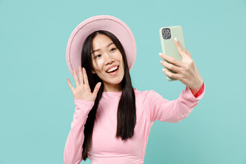 Happy smiling young brunette asian woman 20s wears pink clothes do selfie shot on mobile phone post photo on social network greet waving hand isolated on pastel blue color background studio portrait