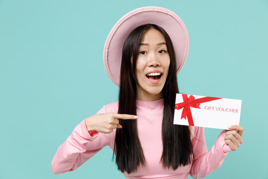 Pleased smiling young brunette asian woman 20s open mouth wear pink clothes hold gift certificate coupon voucher card for store pointing on it isolated on pastel blue color background studio portrait