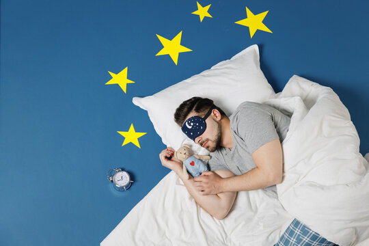 Top view young man in pajamas jam sleep mask resting relax at home lies wrap covered under blanket duvet hug teddy bear plush toy isolated on dark blue sky background. Good mood night bedtime concept.