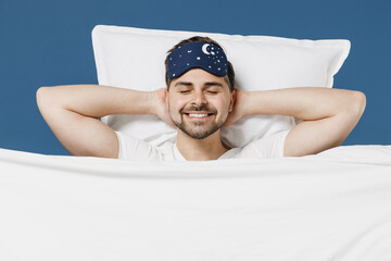 Young man in pajamas jam sleep mask rest relaxing home with lies wrap covered under blanket duvet with closed eyes hands behind neck isolated on dark blue background. Good mood night bedtime concept