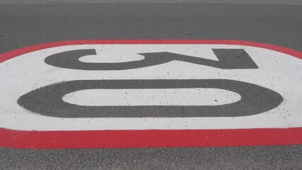 A 30 km per h horizontal speed limit sign painted on an asphalt road. A car driving on the road, which is painted on red, white and gray colours.