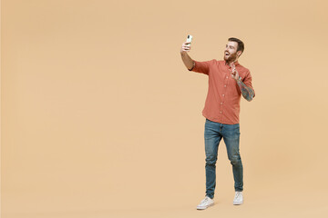 Full size body length fun tatooed young brunet man 20s short haircut wears apricot shirt doing selfie shot on mobile cell phone show victory sign isolated on pastel orange background studio portrait