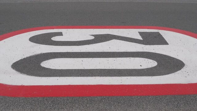A 30 km per h horizontal speed limit sign painted on an asphalt road. A car driving on the road, which is painted on red, white and gray colours.