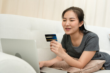 Asian woman holding a credit card for online shopping while sitting on the couch while on vacation at home.