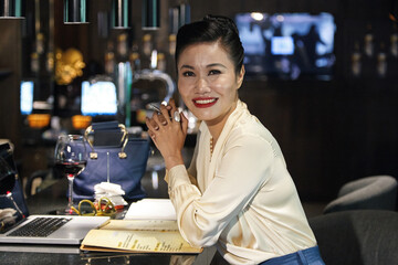Beautful smiling female restaurant manager working on new menu, she is sitting at bar counter and working on laptop