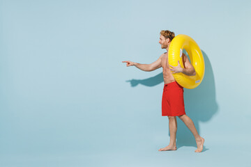 Full length side view young man in red shorts swimsuit near hotel pool hold inflatable ring point finger aside on workspace isolated on pastel blue background Summer vacation sea rest sun tan concept