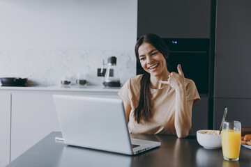 Young housewife woman 20s wear beige shirt eat breakfast use laptop pc computer talk by video call do shaka hand hello gesture cook food in light kitchen at home alone Healthy diet lifestyle concept