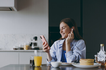 Young housewife woman 20s wear casual clothes blue shirt air pods eat breakfast use mobile cell phone pancakes listen to music cook food in light kitchen at home alone Healthy diet lifestyle concept.