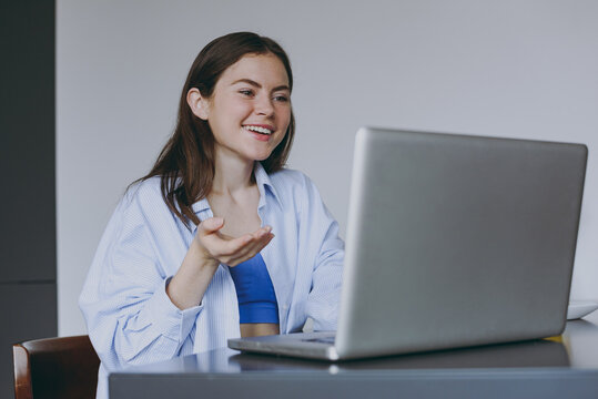 Smiling excited young housewife woman in casual clothes blue shirt talk by video call use laptop pc computer spread hand drink juice cook food in light kitchen at home Healthy diet lifestyle concept.