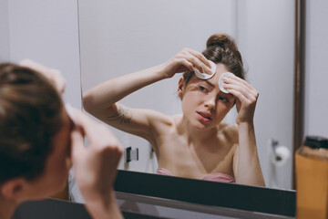 Naklejka premium Disappointed sad half naked young woman 20s wearing bra wipes face with cotton swabs tonic lotion reflected in mirror in bathroom do morning routine Skin care healthcare cosmetic procedures concept..