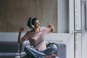 Full length young cheerful fun positive woman in casual clothes headphones sit on grey sofa listen to music new playlist dance rest on weekends indoors flat at home People lifestyle leisure concept
