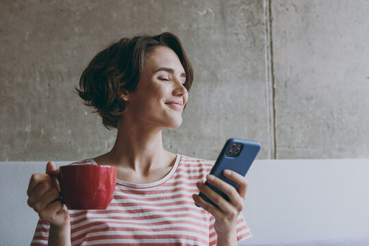 Young smiling satisfied relaxed calm happy 20s wear casual clothes sitting on grey sofa using mobile cell phone hold red cup drink coffee rest indoors flat at home People lifestyle leisure concept