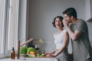 Young amazed fun couple two woman man 20s in casual clothes guy whisper gossip and tells secret behind his hand sharing news cook food in light kitchen at home together Healthy diet lifestyle concept.