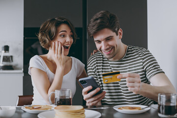 Young amazed couple two woman man in casual clothes sit by table eat pancake use mobile cell phone...