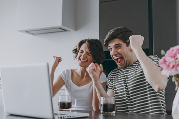 Young couple two woman man fans in casual t-shirt clothes sit table drink coffee watching live stream laptop pc computer do winner gesture in light kitchen at home together People lifestyle concept.