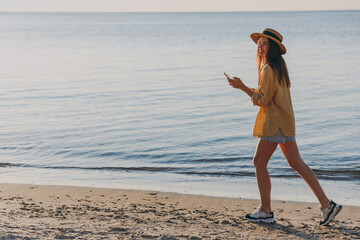 Side profile view full size young woman in straw hat shirt summer clothes walk use modile cell phone outdoors at sunrise sun dawn over sea beach background. People vacation lifestyle journey concept