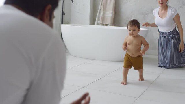 Tracking shot of happy toddler girl walking barefoot from mother to father in bathroom. Baby taking first steps