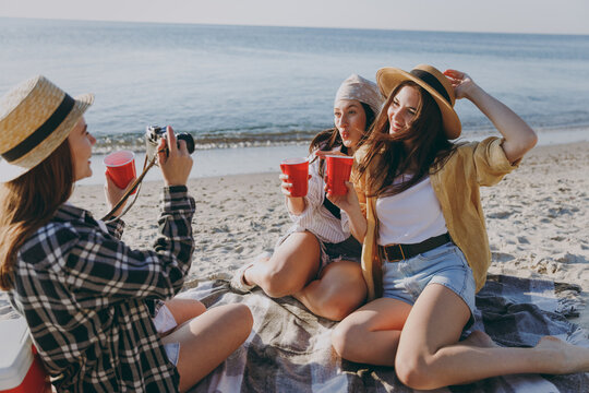 Full body three friends young women 20s in straw hat summer clothes have picnic hang out take photo drink liguor have fun raise toasts outdoors on sea beach background People vacation journey concept.