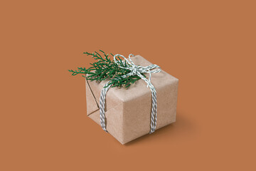 Small Christmas gift box wrapped in craft paper with natural green thuja branch decor isolated on solid beige background. Modern minimal xmas brown backdrop. New Year presents celebration concept 