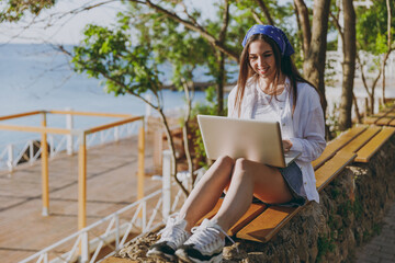 Full length young traveler tourist woman in bandana shirt summer casual clothes rest sit on bench hold use work on laptop pc computer outdoors at sea beach People vacation lifestyle journey concept.