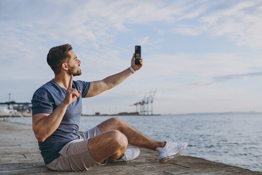 Full body young sporty sportsman man in sports clothes shorts train do selfie shot photo on mobile cell phone show v-sign at sunrise sun over sea beach outdoor on pier seaside in summer day morning.