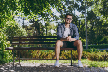 Full body young man in blue shirt shorts glasses sit on bench hold takeaway paper cup drink coffee rest relax in spring green city park sunshine lawn outdoor on nature Urban lifestyle leisure concept