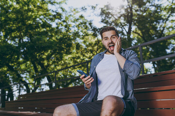 Bottom view young shocked man 20s in blue shirt shorts sitting on bench use mobile phone holding face rest relax in spring green city sunshine park outdoor on nature Urban leisure lifestyle concept.