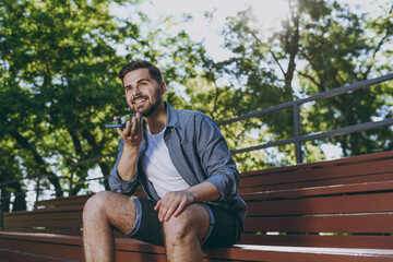 Bottom view young smiling man 20s wear blue shirt shorts sit on bench record voice message on mobile cell phone rest relax in spring green city park outdoor on nature Urban leisure lifestyle concept.