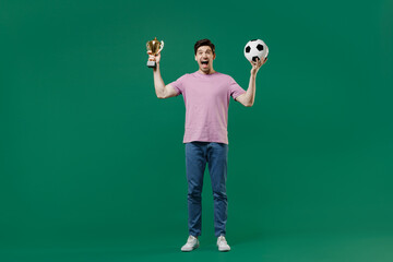 Full size body length young fun man fan wears pink t-shirt cheer up support football sport team hold in hand soccer ball cup watch tv live stream scream isolated on dark green color background studio.