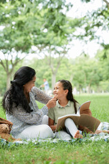 Happy Asian mother sitting on the grass with her daughter on a picnic, they reading a book together
