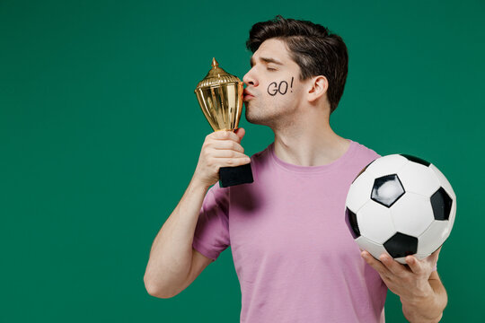Young fun man fan wear basic pink t-shirt cheer up support football sport team hold in hand soccer ball cup champion win cup watch tv live stream scream isolated on dark green color background studio