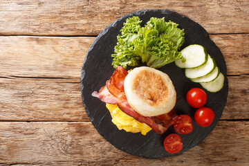 Closeup of a breakfast sandwich with scrambled eggs, bacon and cheese on an english muffin on a slate board on the table. horizontal top view from above