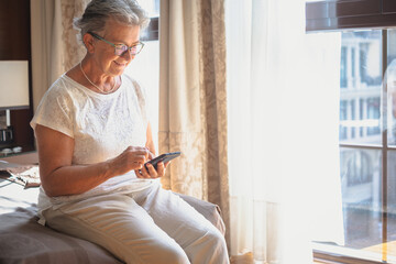 An elderly woman in a hotel room sits on the bed using her mobile phone. A large window behind her