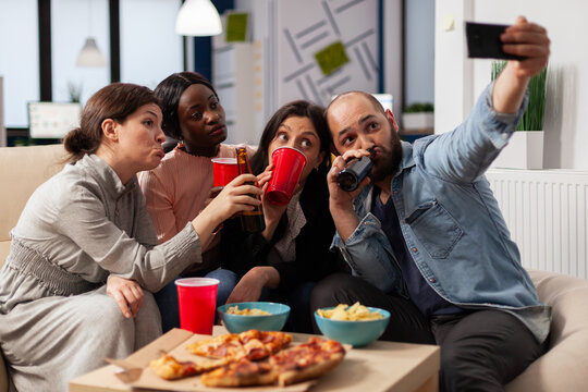 Multi ethnic team of friends make memories at after work party taking pictures on smartphone. Coworkers celebrating at office space with snacks pizza chips cups of drinks on table