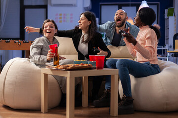 Multi ethnic friends celebrate together after work at office while playing game on tv console with controller joysticks. Diverse group of colleagues having fun gaming at party celebration