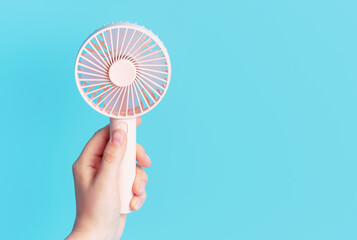 Female hand holding a pink mini fan on blue background close-up.