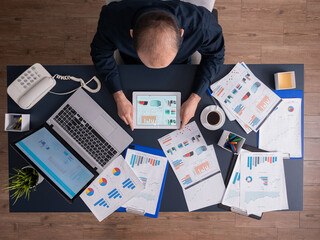 Top view of businessman using tablet pc analyzing financial charts and documents, sitting at desk in corporate office. Employee scrolling through graphs. Successful entrepreneur analyzing strategy.