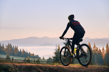 Fototapeta na wymiar Silhouette of cyclist riding bike with coniferous trees and hills on background. Back view of man bicyclist enjoying bicycle ride in mountains in the morning. Concept of biking and active leisure.