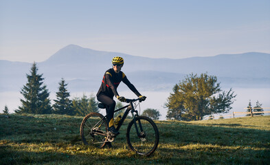 Obraz na płótnie Canvas Smiling man cyclist in cycling suit riding bicycle on grassy hill. Male bicyclist in safety helmet enjoying the view of majestic mountains during bicycle ride. Concept of sport, bicycling and nature.
