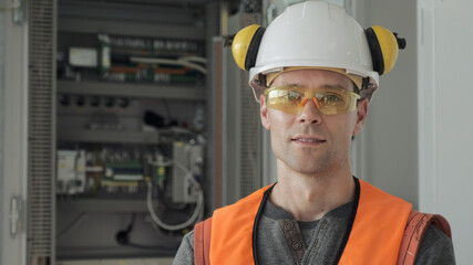 Portrait of young worker wearing helmet and goggles in headphones standing near open switchboard in workshop of production plant close view
