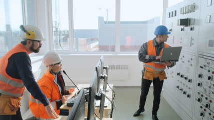 Group of skilled employees in orange uniform and helmets checks operating equipment data with computers in control office of production plant