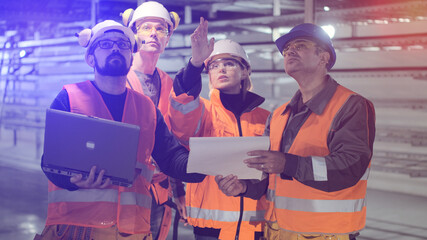 Young woman in orange uniform and helmet with skilled colleagues holding laptop and papers checks workshop building premise at plant