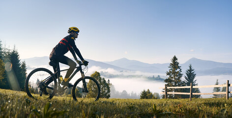 Man riding bicycle on grassy hill and looking at beautiful misty mountains. Male bicyclist enjoying panoramic view of majestic mountains during bicycle ride. Concept of sport, bicycling and nature.