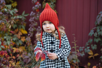 Little toddler girl in red knitted hat standing in autumn scenery