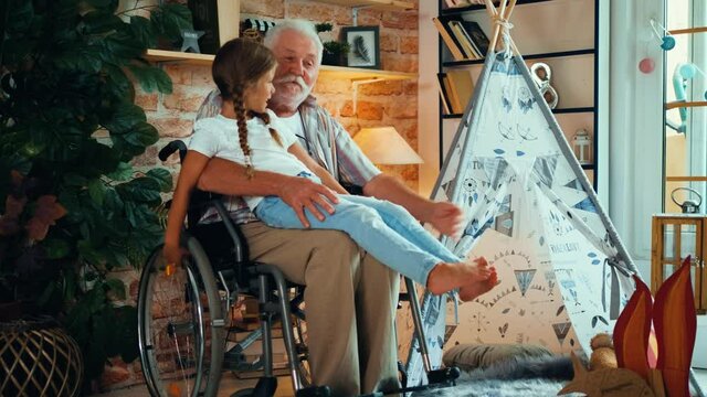 Disabled old man in a wheelchair holds his little granddaughter in his lap. The happy girl listens to her grandfather's stories