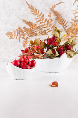 Autumn home decoration with red dried leaves rosehip, fern leaf, ripe berries hawthorn in bowl on white wood table, vertical.