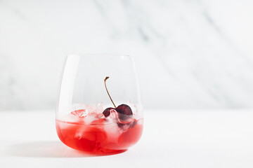 Fresh summer cold cherry alcohol cocktail with ice cubes, slice fruit in elegant white kitchen interior with soft light marble wall.